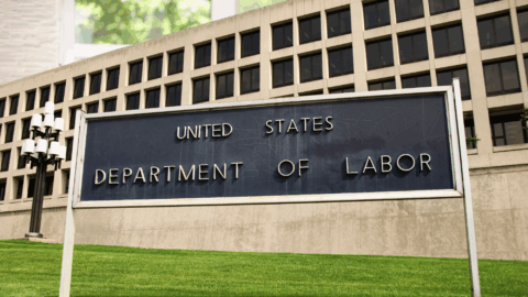 A large sign stands on a green lawn in front of a tall government building. The sign reads “United States Department of Labor.”