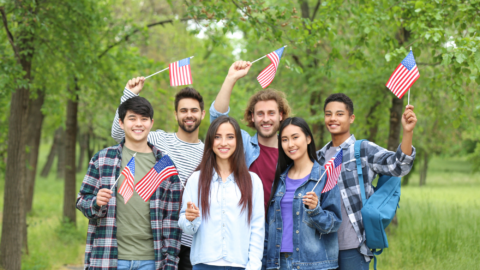 A group of young people is standing outdoors in a green, tree‑filled park, smiling and holding small American flags. They look cheerful and appear to be celebrating or enjoying a patriotic event together.