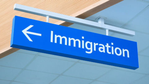 Lentini Visas | Immigration Services, Law Office of Jacqueline Lentini LLC | E-Verify Update: Managing Point of Contact (POC) Information A bright blue airport sign points left toward the immigration area. The sign hangs from the ceiling in a modern terminal.