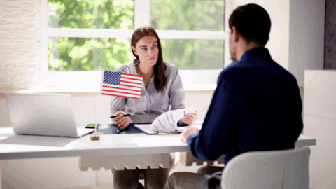 Lentini Visas | Immigration Services, Law Office of Jacqueline Lentini LLC | Good Moral Character & Neighborhood Checks for Naturalization Woman sitting at a desk that has an American flag on it. Man sitting on opposite of the desk.