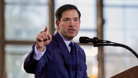 US Secretary of the State Marco Rubio in a suit, speaking at a podium with a strong, focused expression. He raises his hand while talking into the microphone during an indoor event.