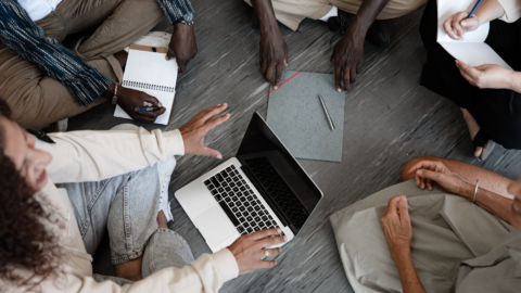 A group of people sit on the floor with laptops. They work together on their computers.