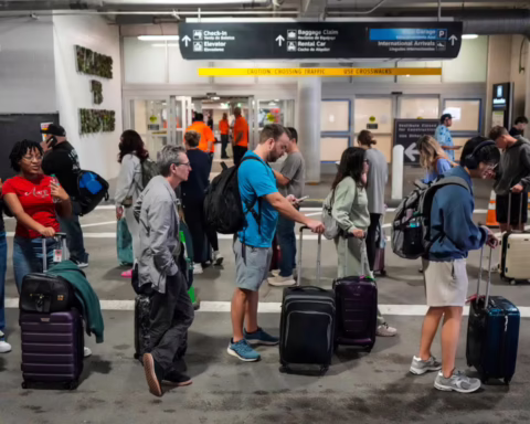 A group of travelers is standing in a line with their luggage outside an airport entrance. The scene looks busy, with people waiting and moving toward the doors.