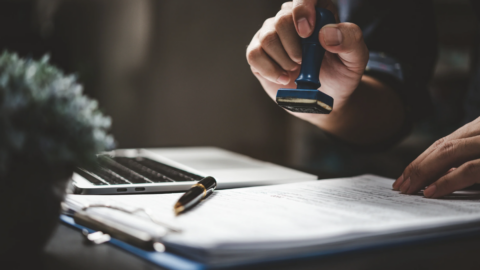 Someone is pressing a rubber stamp onto a document on a desk. A laptop and pen sit nearby, giving the scene a work‑focused feel.