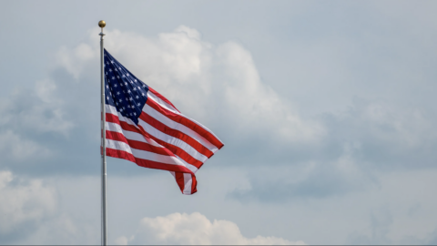 A United States flag waving on a tall pole. Soft clouds fill the sky behind it.