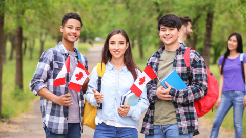 It shows a group of young people outdoors holding small Canadian flags. They look cheerful and appear to be walking together in a green, tree‑filled area.