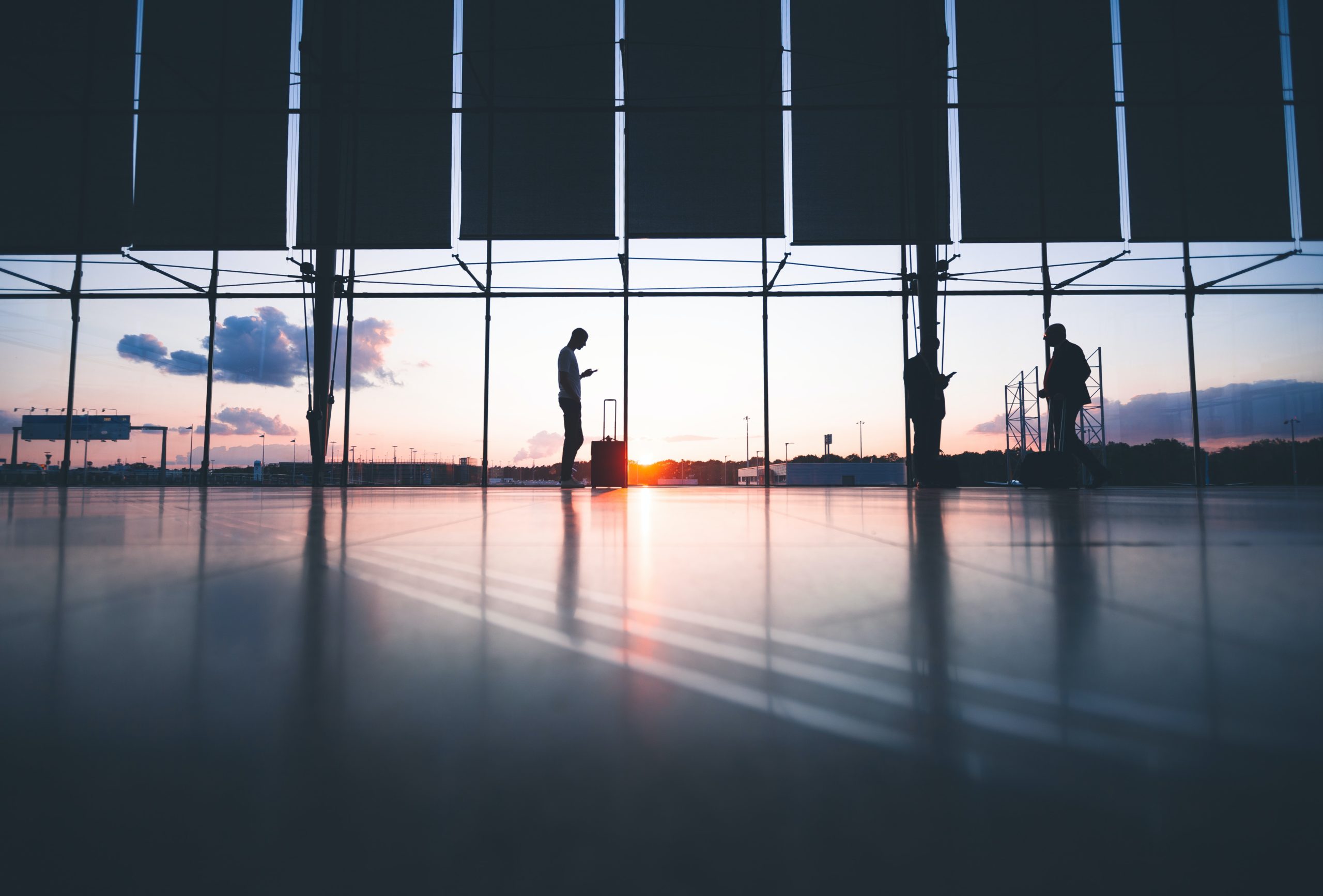 A wide shot of a view from an airport, the sun is setting on the horizon. A silhouette stands in the middle checking their phone.