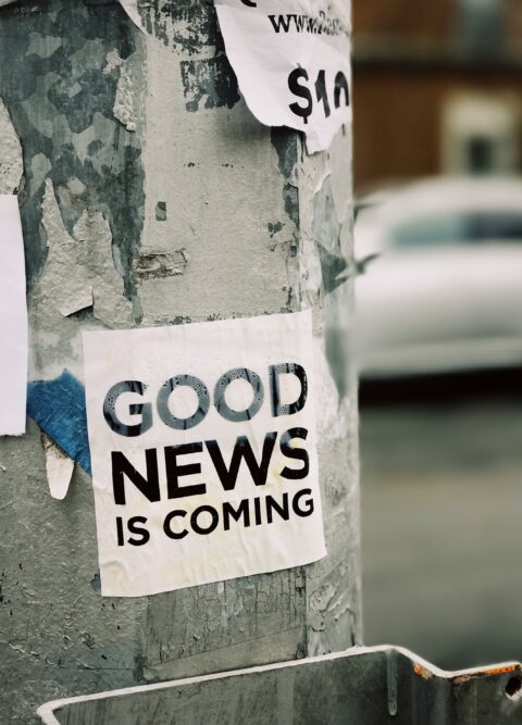 The words "good news is coming" written in black capital letters on a white flyer stuck to a a street post 