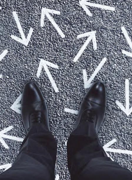 A photograph angled downwards to show business shoes standing on black-top ground with arrows pointing in various directions.