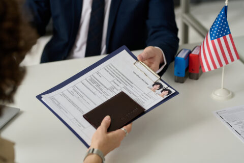 Lentini Visas | Immigration Services, Law Office of Jacqueline Lentini LLC | USCIS Changes H-1B Visa Selection Process A person is handing over a clipboard with forms and a passport to someone across a desk. There’s a small American flag and office supplies on the table.