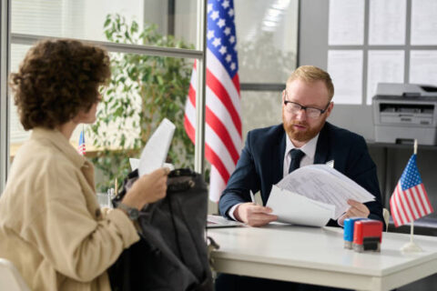 A person gives documents to a man sitting at a desk. The man is checking the papers.