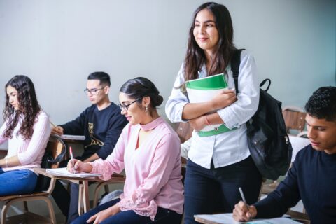 A female student stands by other students sitting in desks.