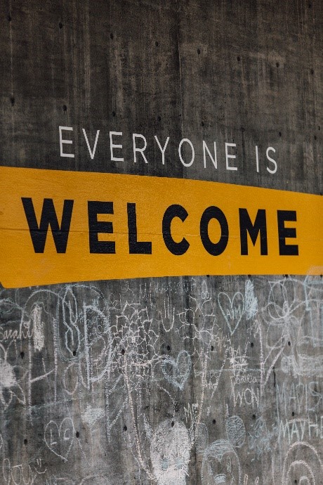 Painted on a concrete wall are the words "Everyone is Welcome." There is a yellow background behind the word welcome. Chalk doodles are drawn underneath the sign. 